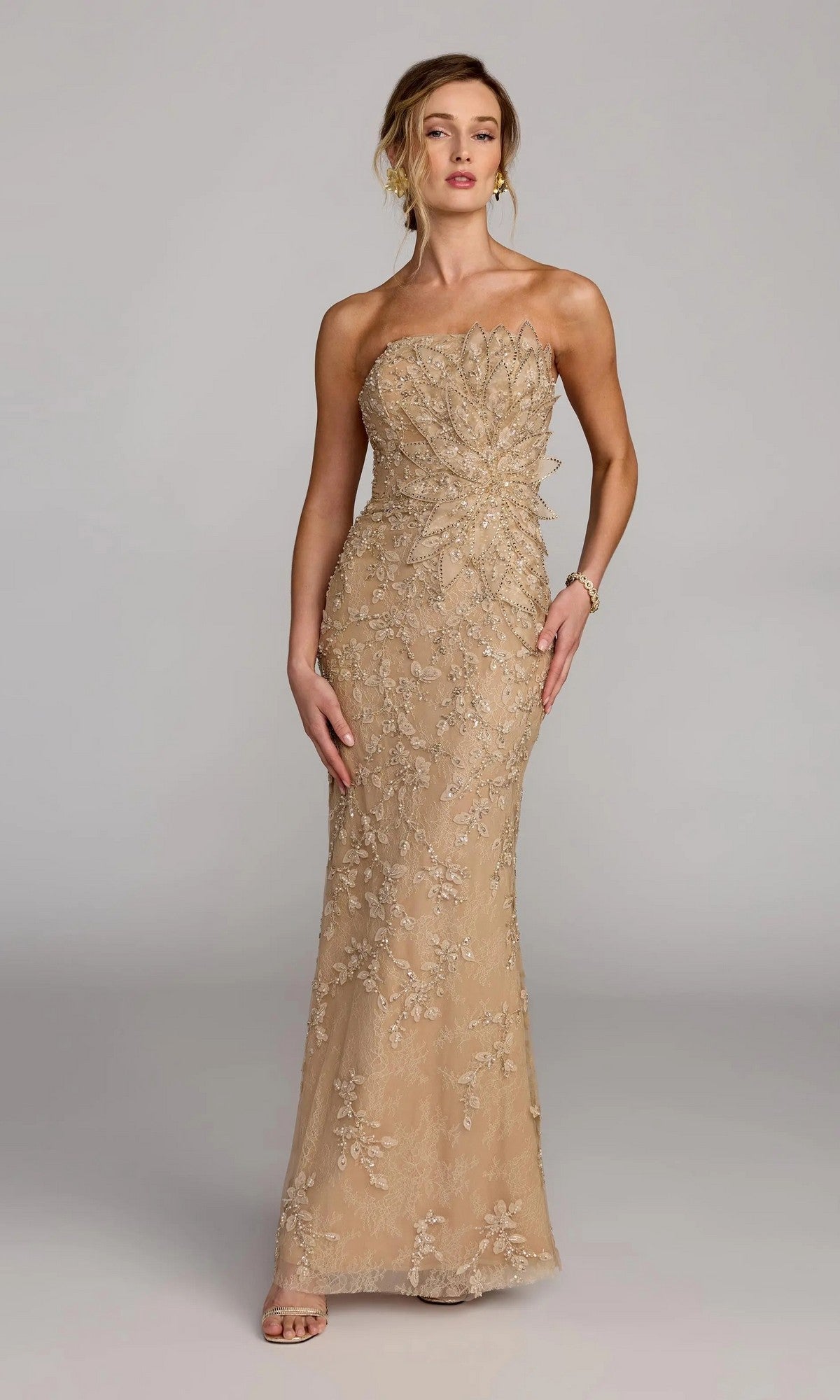A woman wears the Mac Duggal 21200 Long Formal Dress, a gold strapless gown with floral embroidery and a leaf-inspired bodice, standing against a plain background.